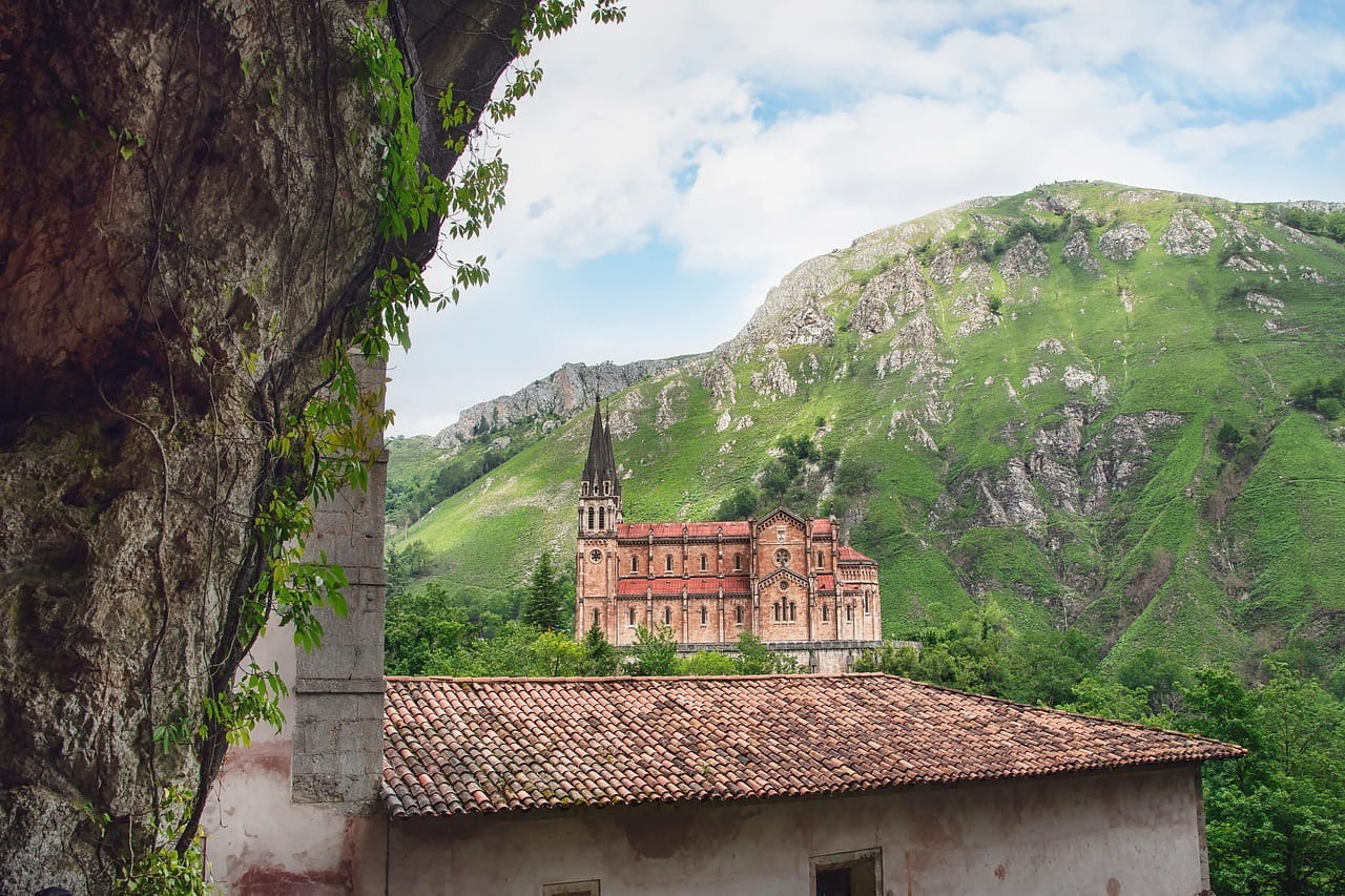 Iglesia histórica de ladrillo rojo con techos de tejas en un valle montañoso verde, visto detrás de un primer plano rocoso y con vegetación en Covadonga, España.