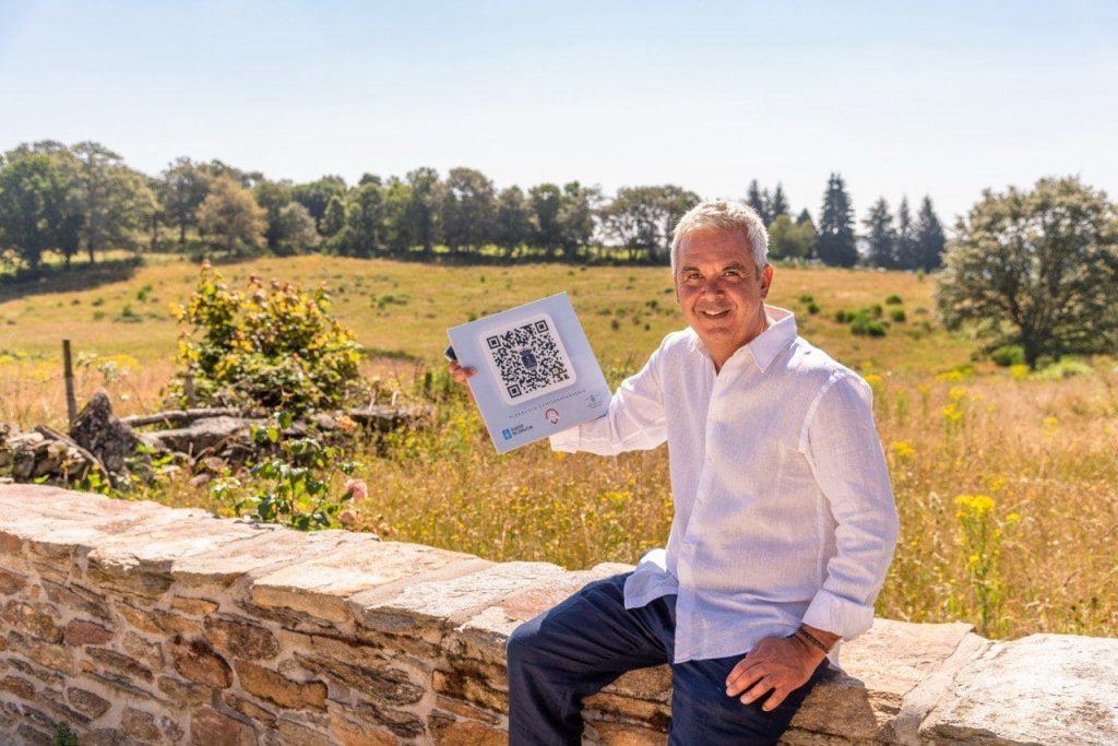 Juan Carlos Pérez, CEO de Aldealista sentado en un muro bajo y mostrando un QR en una baldosa y de fondo un campo rural.