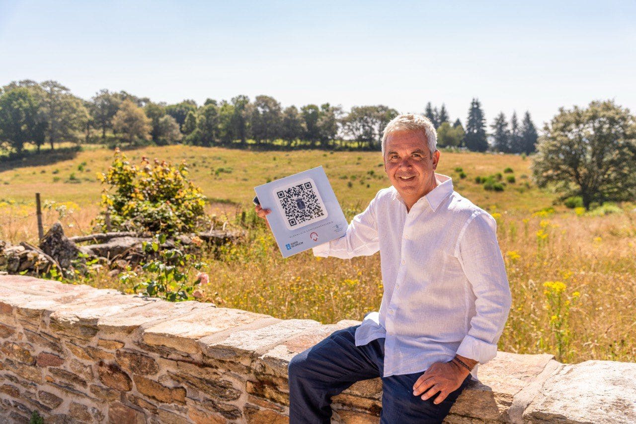 Juan Carlos Pérez, CEO de Aldealista sentado en un muro bajo y mostrando un QR en una baldosa y de fondo un campo rural.