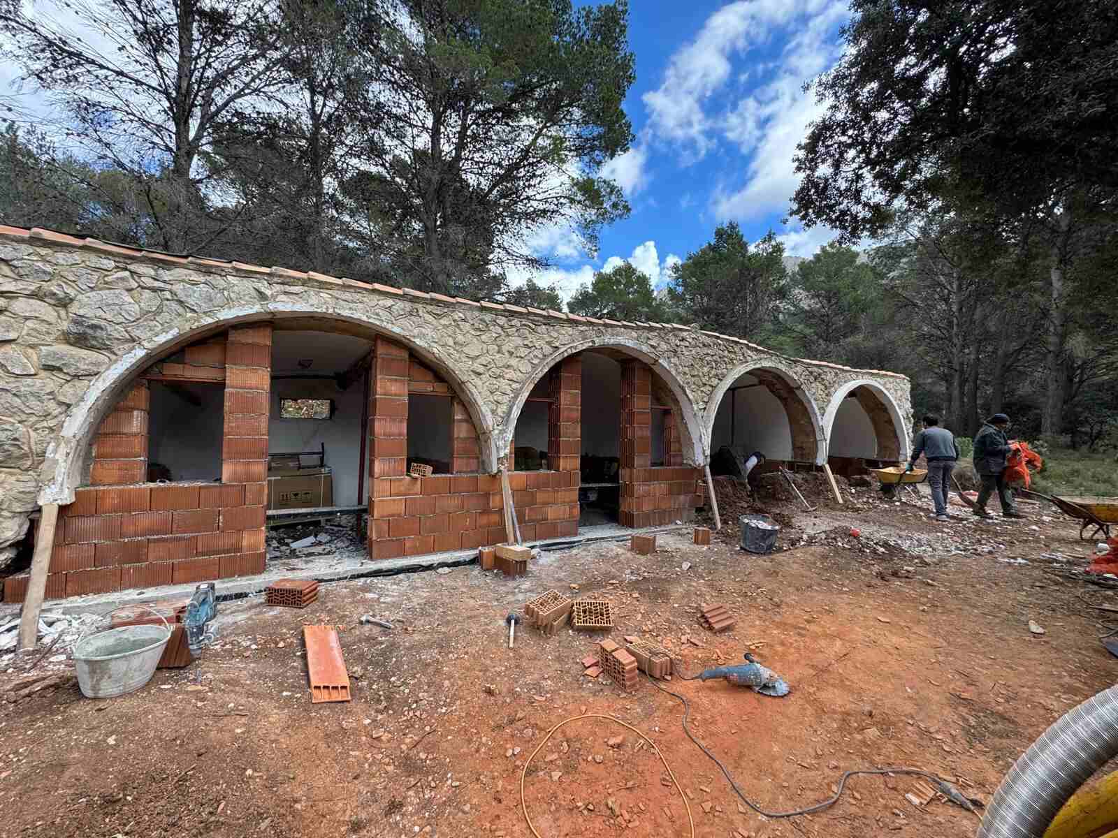Construcción rural de Ruinas Con Futuro en plena rehabilitación, ubicada en un entorno de monte mediterráneo rodeada de pinos. El edificio, de una sola planta, destaca por su fachada formada por cuatro grandes arcos de medio punto en mampostería de piedra vista y rematada con teja cerámica curva, en clara sintonía con la arquitectura vernácula del levante español.