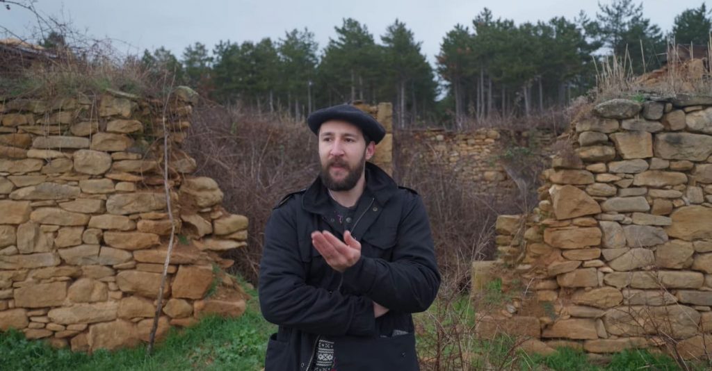 Iker Ganuza (BasqueCraft) habla a cámara con txapela y abrigo negro, de pie entre los muros de piedra en ruinas de una antigua construcción rural, con un bosque de pinos al fondo.