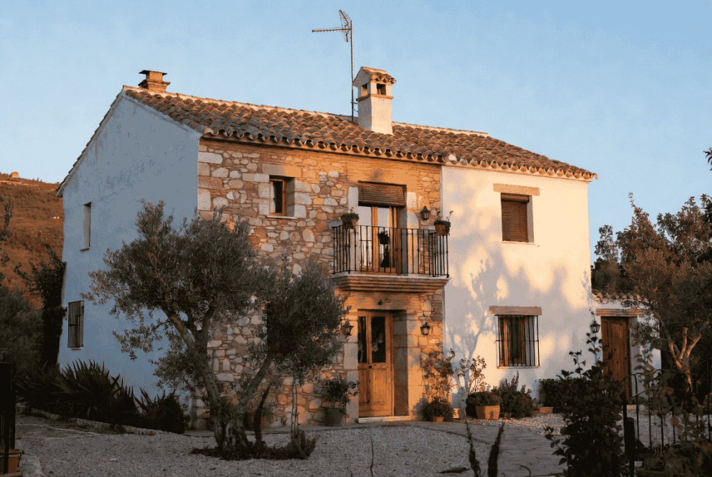Recreación generada con IA de una casa de pueblo rehabilitada, con fachada de piedra y encalada, balcón de forja y tejado de teja, rodeada de olivos al atardecer.