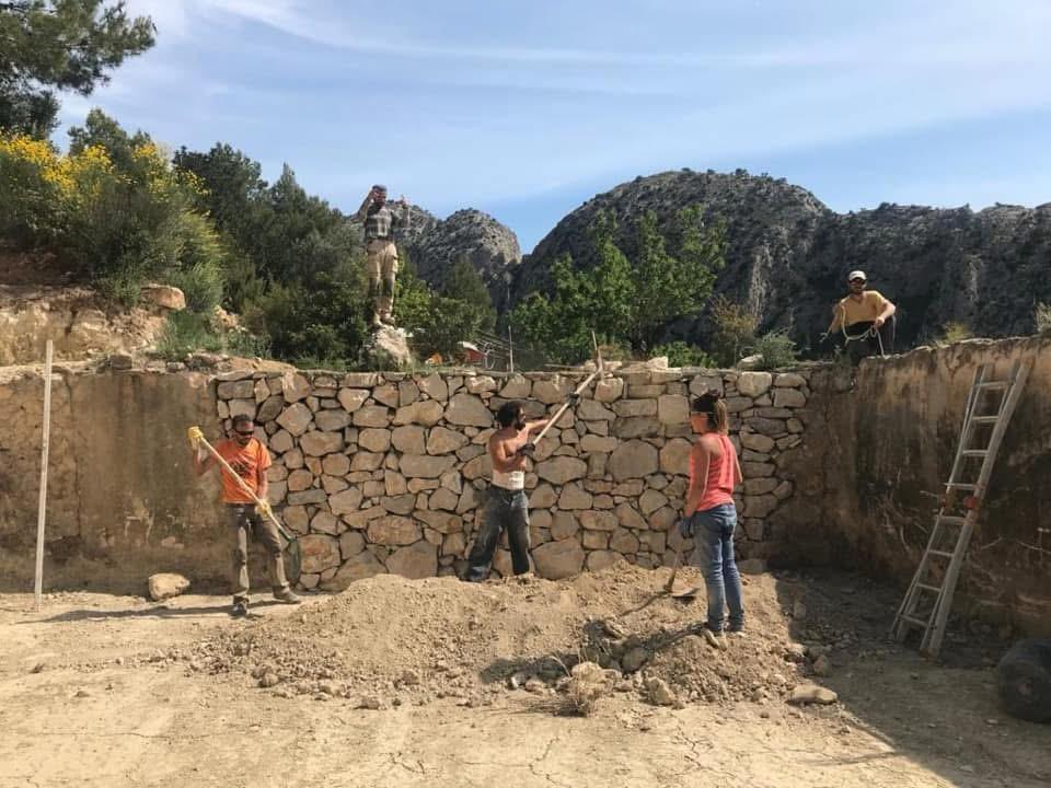 Equipo de la empresa Ruinas con Futuro trabajando en la construcción de un muro de piedra seca en plena reforma rural, con herramientas tradicionales y un paisaje montañoso de fondo.