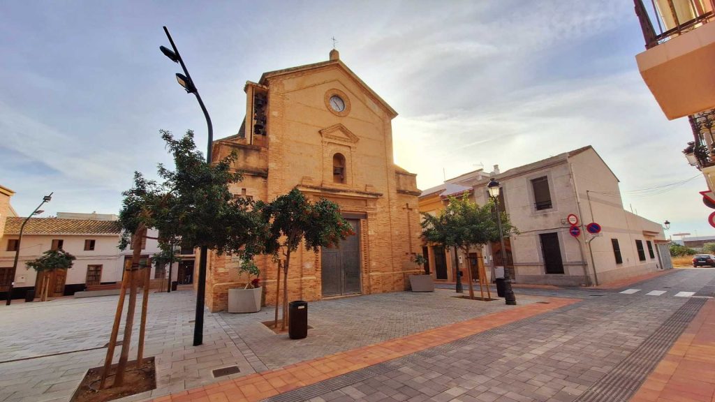 Vista frontal de la iglesia de ladrillo de Llocnou de la Corona, con un reloj en la parte superior y grandes puertas de entrada. Delante de la iglesia hay una plaza peatonal arbolada con farolas y papeleras, flanqueada por casas bajas tradicionales bajo un cielo despejado.