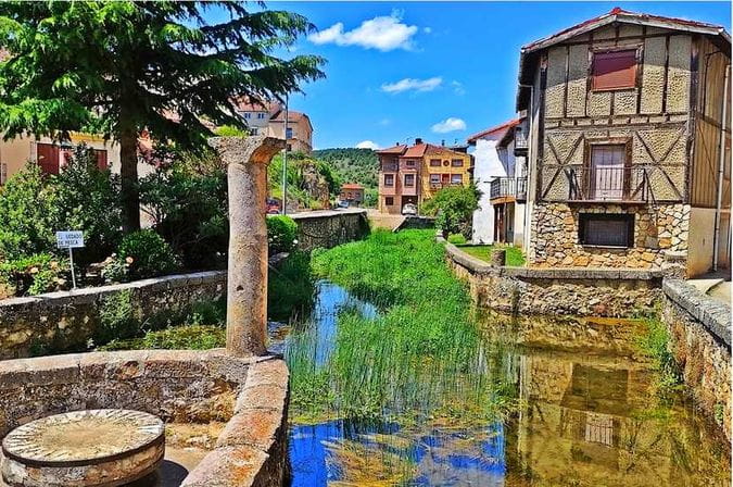 Fotografía a color de Huerta del Rey, Burgos, mostrando el canal de agua central con vegetación, una columna de piedra antigua en primer plano y casas tradicionales con entramado de madera al fondo, bajo un cielo azul con nubes.