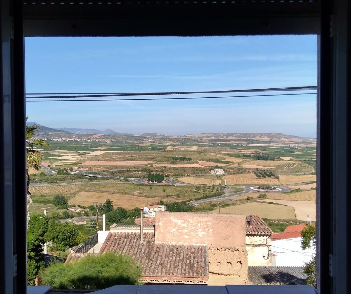 Vista panorámica de un paisaje agrícola desde una ventana, con campos de cultivo, caminos y construcciones rurales en primer plano.
