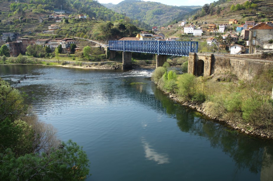 Puente ferroviario azul cruzando el río en Os Peares, Galicia, con casas y colinas verdes al fondo.