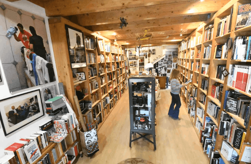Vista interior de la librería Primera Página en Urueña, con estanterías altas de madera repletas de libros a ambos lados, suelo de parquet, una vitrina central con cámaras fotográficas y una persona consultando un libro al fondo.