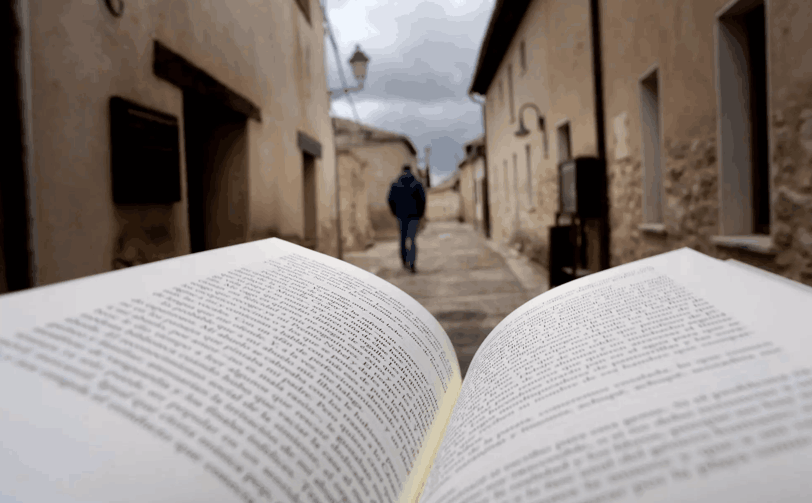 Libro abierto en primer plano en una calle empedrada de Urueña (Valladolid), con casas de piedra a los lados y la figura desenfocada de una persona caminando al fondo.