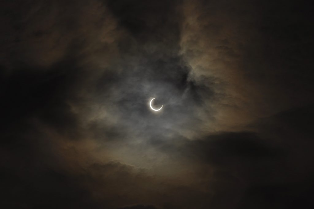 Fase parcial o transitoria de un eclipse solar total en el centro de un cielo oscuro y atmosférico. El sol está cubierto en su mayoría, mostrándose como una brillante y fina silueta en forma de media luna, rodeado de nubes densas de tonos grises y cobrizos iluminadas suavemente por los bordes.