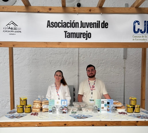 Álvaro Ruiz, presidente de la Asociación Juvenil de Tamurejo, junto a otro miembro en un stand con productos locales, bajo un cartel de la asociación